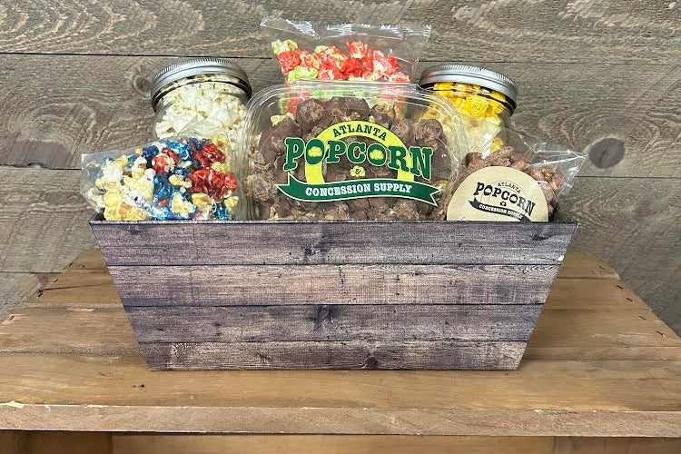 A wooden gift basket filled with assorted popcorn and candy items, including a large jar labeled Atlanta's Popcorn & Concession Supply, on a wooden surface against a rustic wood plank background.