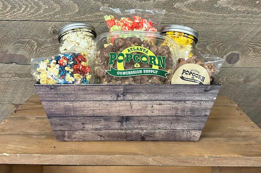 A wooden gift basket filled with assorted popcorn and candy items, including a large jar labeled Atlanta's Popcorn & Concession Supply, on a wooden surface against a rustic wood plank background.